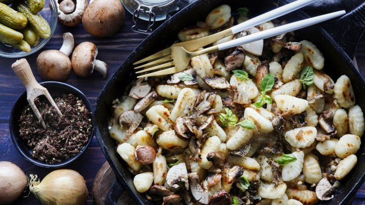 In einer Pfanne liegen frisch zubereitete H&uuml;ttenk&auml;se-Gnocchi mit Pilzen, umgeben von Gew&uuml;rzen und Beilagen.