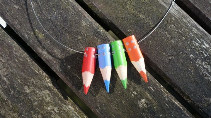 F&uuml;nf geometrische Buntstifte als Schmuck h&auml;ngen an einem Draht, liegend auf einer Holzoberfl&auml;che im Freien.