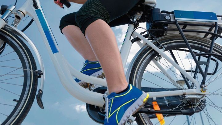 Ein E-Bike f&auml;hrt auf einer sonnigen Strecke, w&auml;hrend eine Person mit sportlichen Schuhen in die Pedale tritt.
