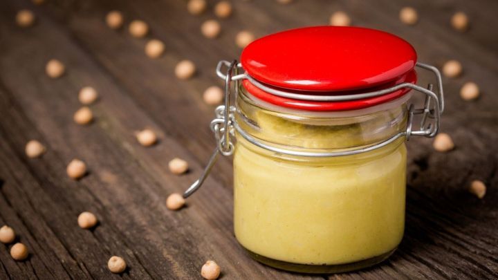 Ein Glas mit rotem Deckel enth&auml;lt einen gelben Dip, umgeben von Senfk&ouml;rnern auf einer rustikalen Holzplatte.