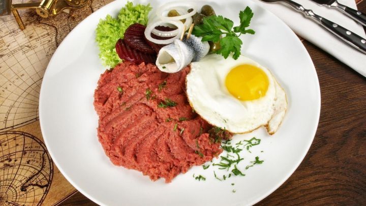 Auf einem Teller liegt Labskaus, eine meist rote Masse, garniert mit Spiegelei, Zwiebeln und Beilagen.
