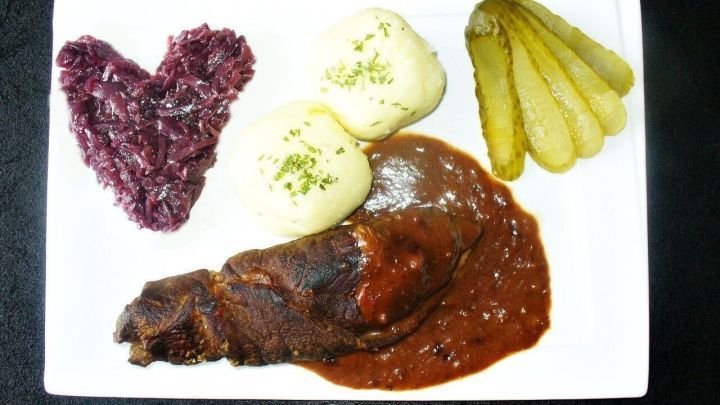 Zarte Rinderroulade liegt in w&uuml;rziger So&szlig;e, umgeben von Kartoffelkn&ouml;deln, Rotkohlherz und Gurken. Ein herzhaftes Gericht.