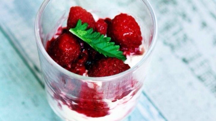 Ein Becher mit Himbeer-Windbeutel-Dessert, gef&uuml;llt mit frischen Himbeeren und einem Spritzer Minze, steht auf einem hellen Holztisch.