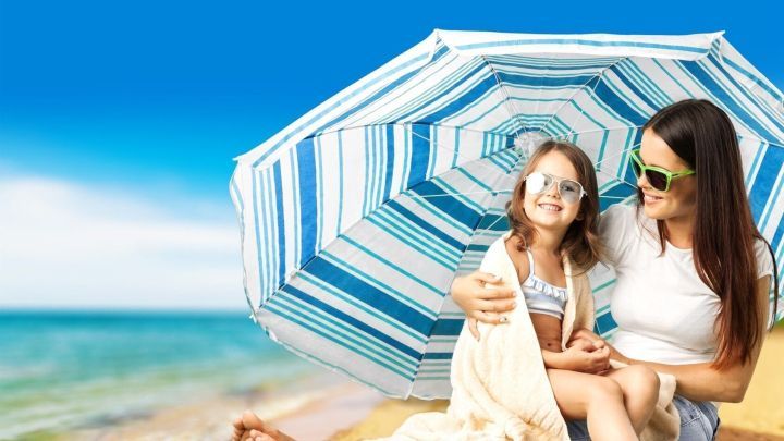 Eine Mutter sitzt mit ihrer Tochter unter einem Sonnenschirm am Strand, um sich vor der Sonne zu sch&uuml;tzen.