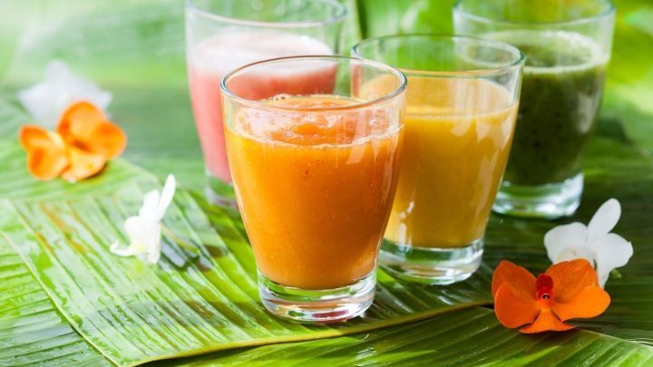 Bunte Smoothies stehen in Gl&auml;sern auf gr&uuml;nen Bl&auml;ttern, umgeben von dekorativen Blumen, die frische gesunde Genussmomente ausstrahlen.