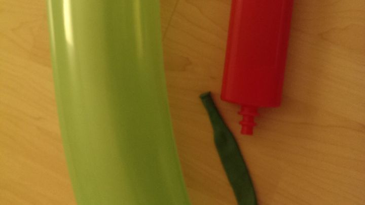 Ein gr&uuml;ner Luftballon in Form einer Gurke liegt neben einer roten, mechanischen Pumpe auf einem hellen Holztisch.