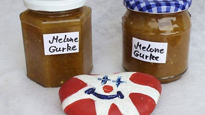 Zwei Gl&auml;ser Melonen-Gurken-Marmelade stehen dekorativ nebeneinander, w&auml;hrend ein bemalter Stein mit einem Clowns-Gesicht die Szene erg&auml;nzt.
