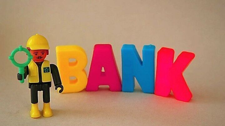 Ein Spielzeugarbeiter steht mit einer Lupe neben bunten Buchstaben, die das Wort "BANK" bilden, auf einem neutralen Hintergrund.