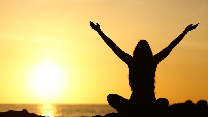 Eine Person sitzt mit erhobenen Armen vor einem Sonnenuntergang am Meer, symbolisiert selbstbestimmtes Leben und Freiheit.