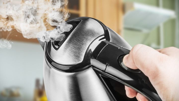 Ein Wasserkocher dampft, w&auml;hrend eine Hand den Knopf dr&uuml;ckt, in einer modernen K&uuml;che mit Holzregalen im Hintergrund.