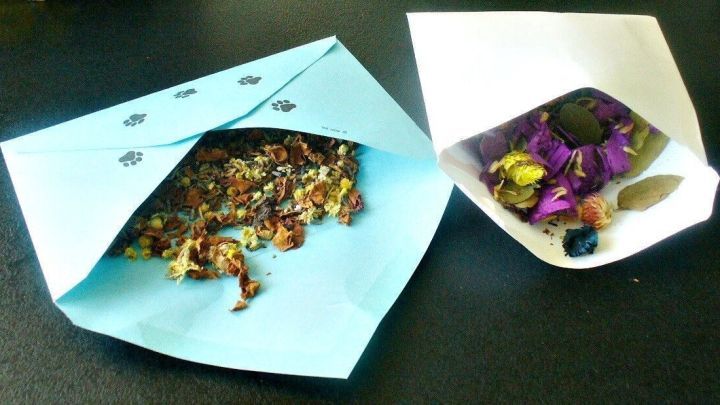 Zwei dekorative Umschl&auml;ge, einer in Himmelblau mit getrockneten Blumen und einer in Wei&szlig; mit bunten Kr&auml;utern, stehen auf einem Tisch.