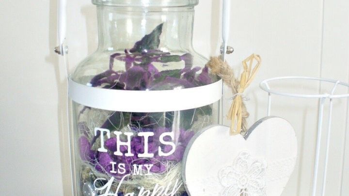 Ein Glas-Windlicht mit lila Dekoration und der Aufschrift &bdquo;THIS IS MY Happy Place&ldquo; steht auf einem Tisch.