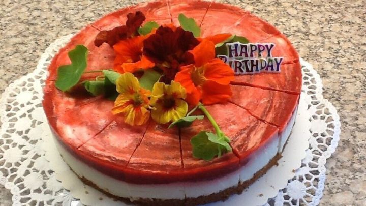 Eine Joghurt-Erdbeer-Torte steht dekoriert auf einem Teller, umgeben von bunten Blumen, mit der Aufschrift &bdquo;Happy Birthday&ldquo;.