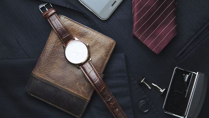 Eine elegante Armbanduhr liegt auf einem braunen Leder-Portemonnaie, umgeben von einer Krawatte und weiteren Accessoires.