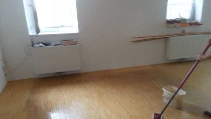 Ein Parkettboden wird mit einem Wischmopp gereinigt, w&auml;hrend Licht durch die Fenster in den Raum f&auml;llt.