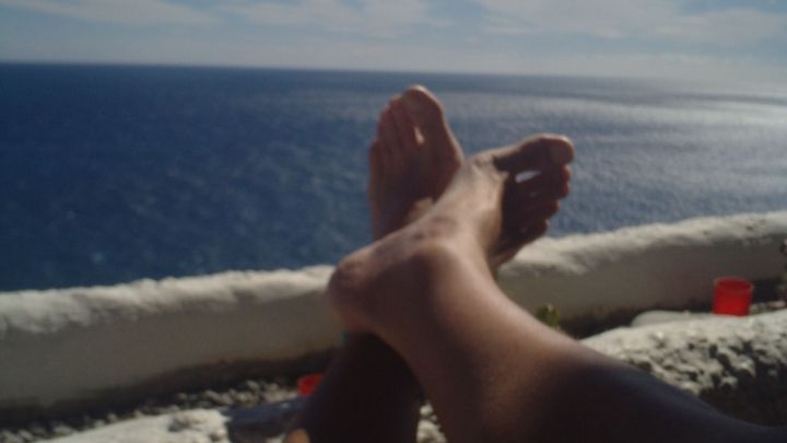 F&uuml;&szlig;e entspannen bequem auf einer wei&szlig;en Mauer, w&auml;hrend der Ozean unter einem sonnigen Himmel im Hintergrund glitzert.