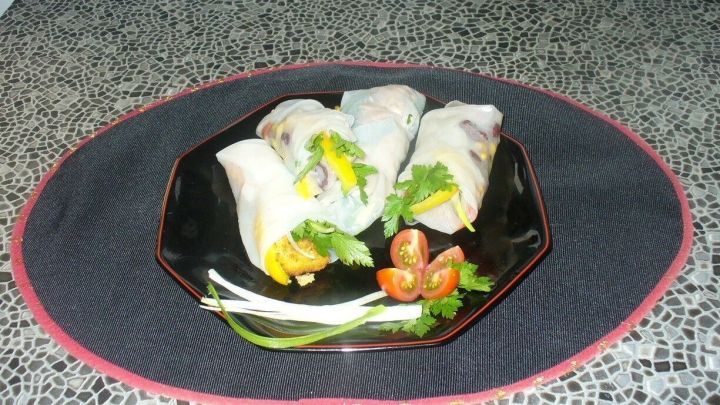 Frische Sommerrollen liegen auf einem schwarzen Teller, umgeben von Kr&auml;utern und Tomaten, appetitlich angerichtet.