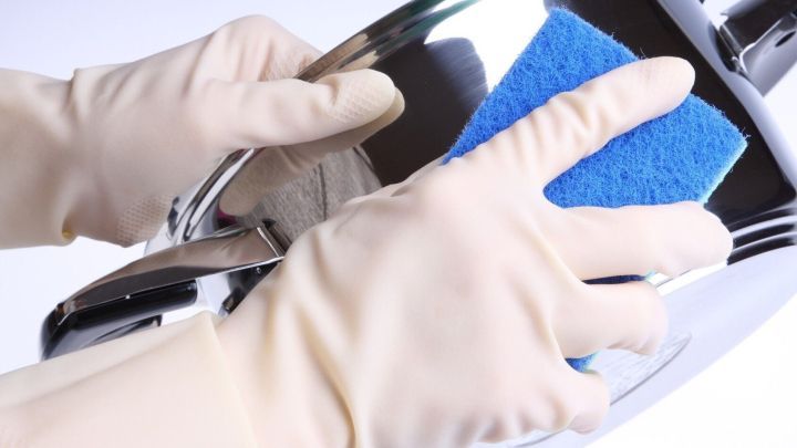 Eine Person mit Gummihandschuhen reinigt mit einem blauen Schwamm fettiges Plastikgeschirr in einer hellen Umgebung.