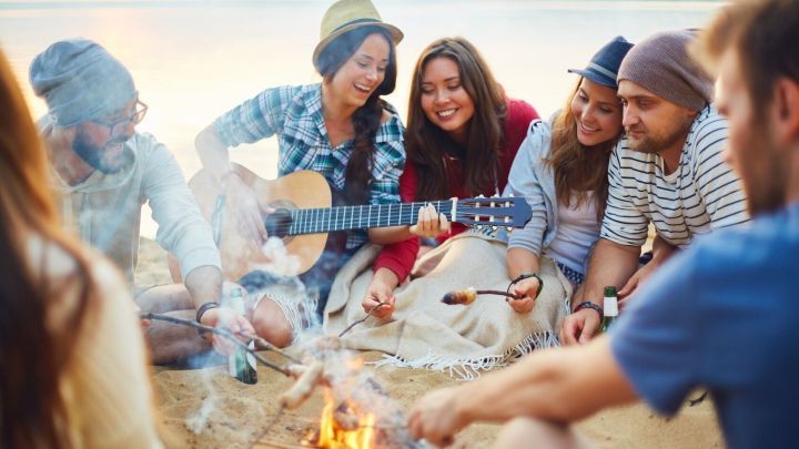 Eine Gruppe junger Menschen sitzt um ein Lagerfeuer, spielt Gitarre und genie&szlig;t z&uuml;nftiges Essen am Strand.