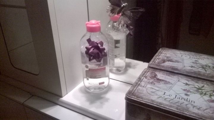 Eine Flasche Mizellen Reinigungswasser steht auf einem wei&szlig;en Waschbecken neben einem dekorativen Beh&auml;lter und einem Blumenstrau&szlig;.