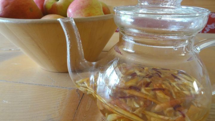 Eine Teekanne mit Apfelschalentee steht auf einem Tisch, w&auml;hrend &Auml;pfel in einer Sch&uuml;ssel im Hintergrund liegen.