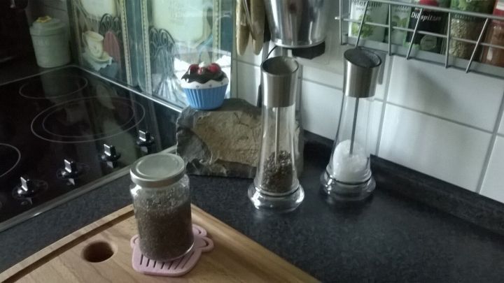 Ein Glas mit Chia-Samen steht auf einem Holzbrett in einer modernen K&uuml;che, umgeben von Gew&uuml;rzm&uuml;hlen und Kochutensilien.