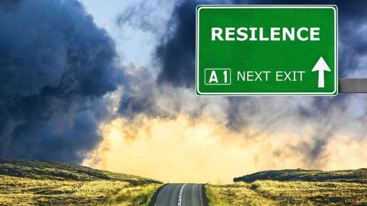 Eine Stra&szlig;e f&uuml;hrt in die Ferne, w&auml;hrend ein Schild "RESILIENCE (A1 NEXT EXIT)" die Richtung anzeigt. Dunkle Wolken h&auml;ngen &uuml;ber der Landschaft.