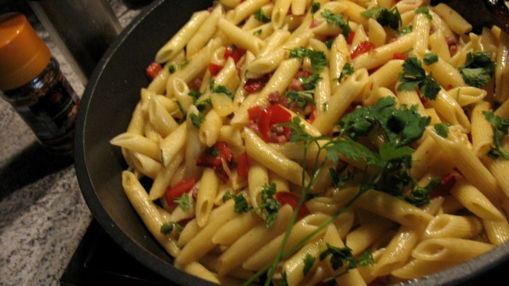 Eine Pfanne mit Penne-Pasta, Paprika und frischen Kr&auml;utern wird in einer dunklen K&uuml;che zubereitet.