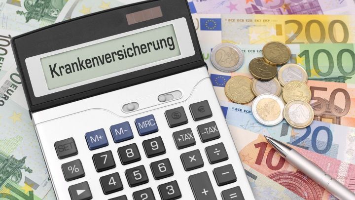 Ein Taschenrechner zeigt "Krankenversicherung" an, umgeben von Euro-M&uuml;nzen und -Scheinen, die finanzielle Aspekte verdeutlichen.