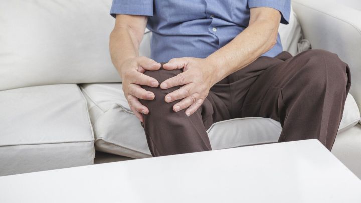 Ein Mann sitzt auf einem Sofa und h&auml;lt sich schmerzhaft das Knie, w&auml;hrend er sich offensichtlich unwohl f&uuml;hlt.