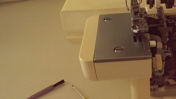 Eine Overlockmaschine steht bereit, w&auml;hrend eine Person Nadeln wechselt. Werkzeuge wie Schraubendreher und Nadeln liegen daneben.