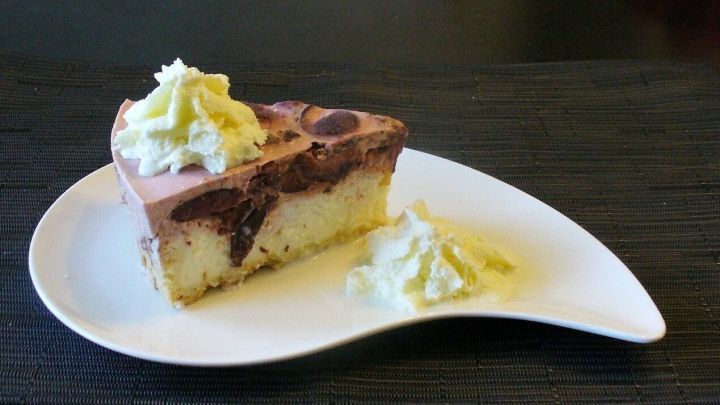 Ein St&uuml;ck K&auml;sekuchen mit einer cremigen Pflaumen-Frischk&auml;setopping ist auf einem eleganten wei&szlig;en Teller angerichtet und umgeben von einem schlichten Schwarz.