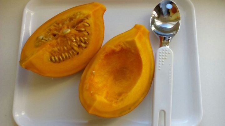 Ein halber K&uuml;rbis liegt auf einem wei&szlig;en Teller, ausgeh&ouml;hlt mit einem L&ouml;ffel daneben, bereit zum Entkernen.