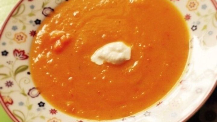 Eine Sch&uuml;ssel K&uuml;rbiscremesuppe mit einem Klecks Sahne steht auf einem bunten Teller auf einem gr&uuml;nen Untergrund.