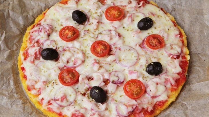 Eine runde glutenfreie Polenta Pizza liegt auf Backpapier, belegt mit Tomaten, Oliven und K&auml;se, frisch gebacken.