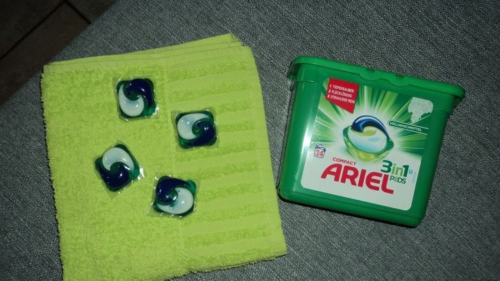 Vier Ariel 3in1 Pods liegen auf einem gr&uuml;nen Tuch, neben einer Verpackung in einem neutralen Umfeld.