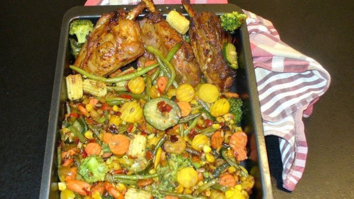 Saftige Lammhaxen liegen neben buntem Ofengem&uuml;se in einer Backform, umgeben von einem gestreiften K&uuml;chentuch.