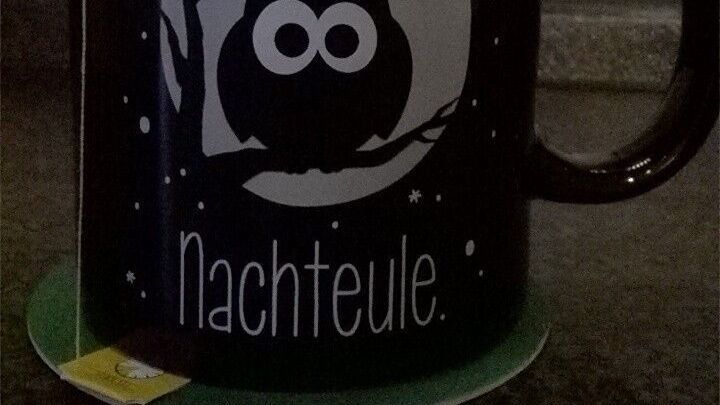Eine schwarze Tasse mit einem Eulenmotiv und dem Schriftzug "Nachteule" steht auf einem Untersetzer in einer dunklen K&uuml;che.