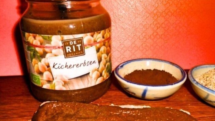 Ein Glas Schoko-Hummus steht neben Sch&uuml;sseln mit Kakao und Sesam auf einem Tisch mit einem Holzbrett im Hintergrund.