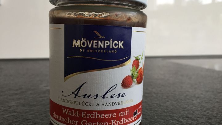 Ein Marmeladenglas steht auf einem Tisch, w&auml;hrend der Besitzer es mit einem Spachtel von Kleberesten auf Glas reinigt.