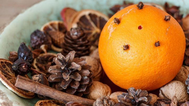 Eine Apfelsine mit Nelken steht im Mittelpunkt einer dekorativen Schale, umgeben von Tannenzapfen und getrockneten Fr&uuml;chten.