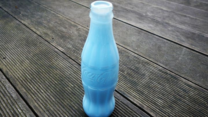 Eine bunte Colaflasche steht auf einem Holzfu&szlig;boden und dient als Vase, kreativ umfunktioniert f&uuml;r Blumen.