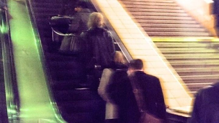 Eine Gruppe von Menschen mit Rucks&auml;cken benutzt die Rolltreppe in einem gesch&auml;ftigen, beleuchteten Bahnhof.