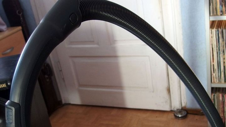 Der Staubsaugerschlauch liegt gebogen auf einem Bodenbelag, w&auml;hrend der Raum mit einem Regal und einer T&uuml;r im Hintergrund endet.