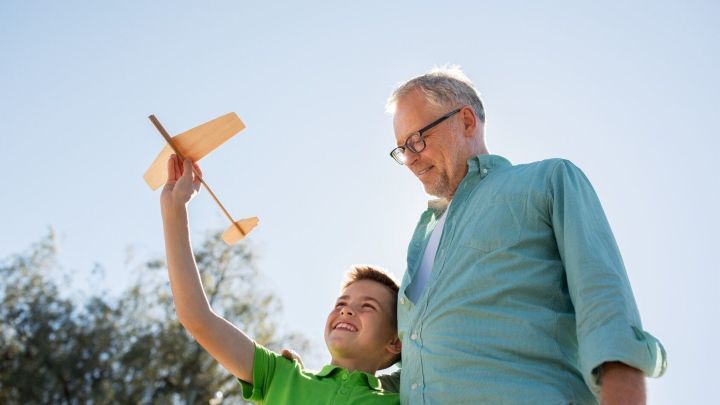 Ein Junge hebt stolz ein Holzflugzeug in die Luft, w&auml;hrend er neben einem &auml;lteren Mann im Freien steht, beide l&auml;chelnd.
