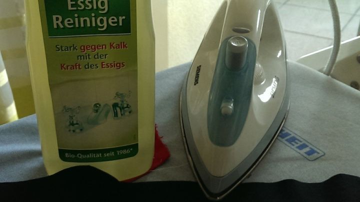 Ein Essigreiniger steht neben einem B&uuml;geleisen auf einem Hemd, w&auml;hrend ein B&uuml;gelvorgang in einer hellen Umgebung stattfindet.