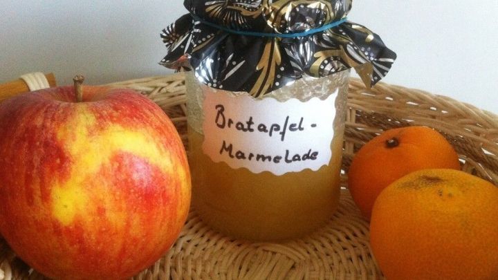 Ein Glas Bratapfelmarmelade steht im Korb, umgeben von einem roten Apfel und einer orangefarbenen Frucht, in einem hellen Raum.