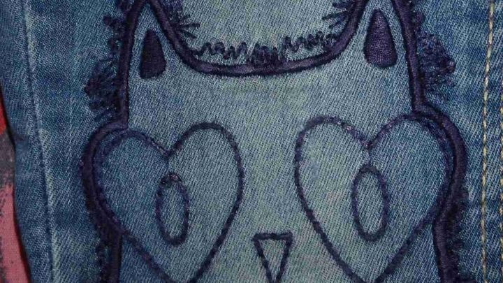 Eine Jeans mit einem niedlichen Katzenmotiv auf der Tasche, die zum Flicken vorbereitet ist.