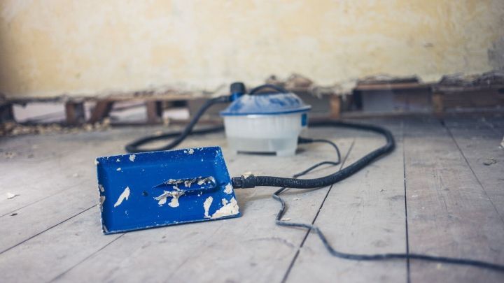 Ein blauer Styroporkleber-Entferner liegt auf dem Boden, w&auml;hrend er eine Wand in einem renovierungsbed&uuml;rftigen Raum bearbeitet.