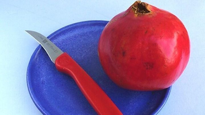 Ein Granatapfel liegt auf einem blauen Teller neben einem roten Messer, bereit zum Entkernen ohne Spritzer.